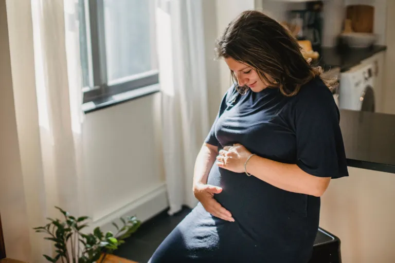 Femme enceinte en robe bleu marine, les mains sur son ventre, près d'une fenêtre