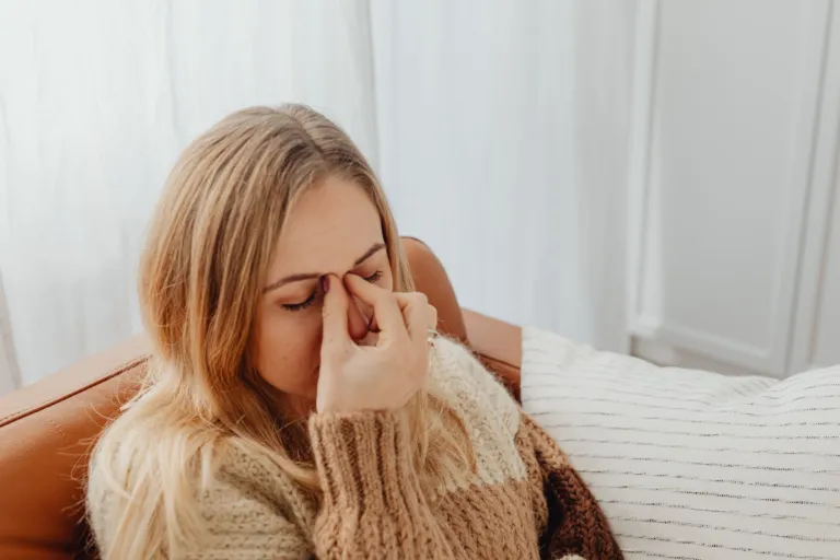 Femme blonde assise sur un canapé, se pinçant l'arête du nez, yeux fermés